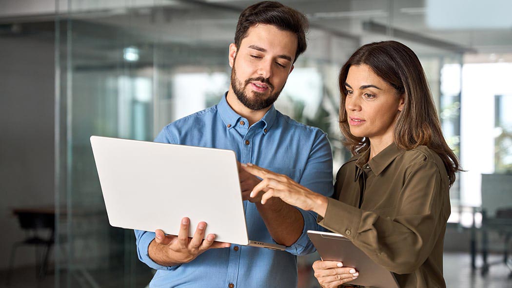 Deux professionnels debout dans un bureau moderne, collaborant en regardant un ordinateur portable et tenant une tablette, avec un bouton lecture indiquant qu’il est possible de cliquer sur l’image pour lancer une vidéo