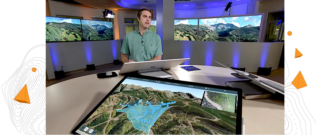 Homme devant une grande table ronde sur laquelle se trouve un grand écran d’ordinateur et, derrière lui, deux écrans de télévision grand format montrant une image 3D numérique