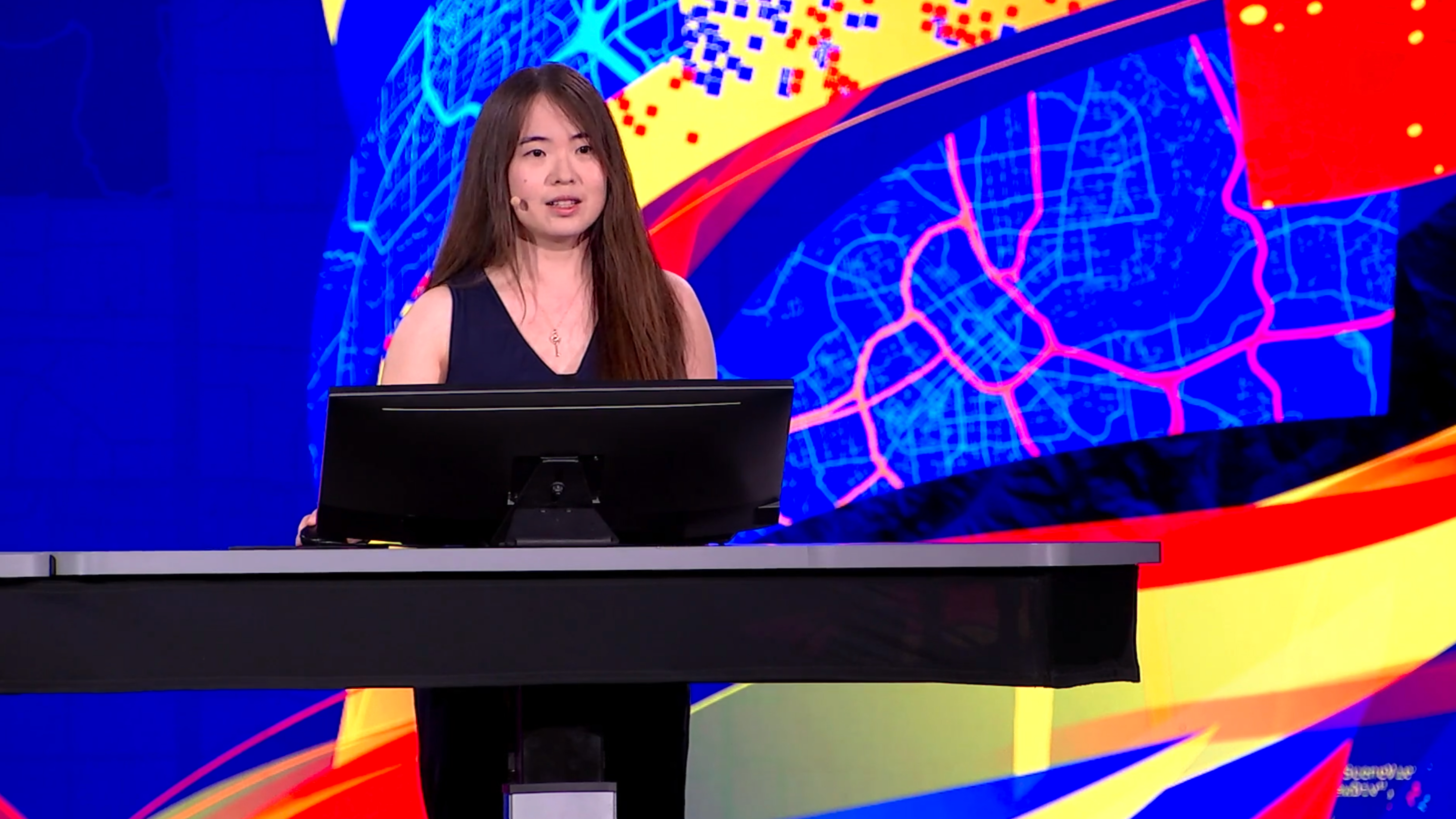 Jeune femme en robe bleue, debout derrière un pupitre, avec projection de carte d’une ville derrière elle