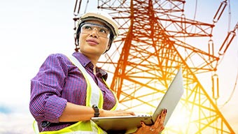 Ouvrière de chantier portant une veste jaune avec un ordinateur portable ouvert devant un pylône électrique