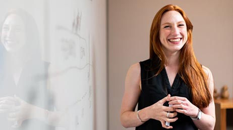 Personne souriante portant un haut noir sans manches et se tenant debout à côté d’un tableau de présentation dans un cadre professionnel décontracté