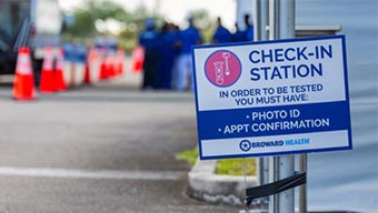 Rue où sont alignés des cônes orange et groupe de travailleurs portant des shorts bleus à côté d’un panneau indiquant un poste d’enregistrement