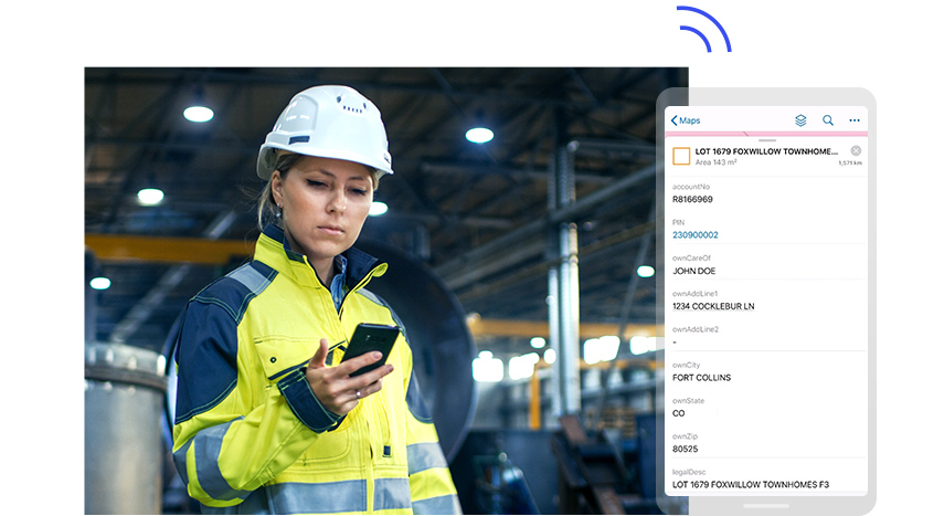 Personne portant un casque de sécurité blanc et une veste de sécurité jaune consultant un téléphone portable qu’elle tient à la main, à côté d’un grand écran de téléphone portable affichant les données d’un compte personnel fictif
