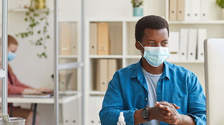 Jeune homme portant un masque chirurgical bleu, assis devant son ordinateur dans un bureau