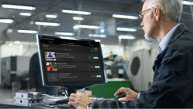 Homme portant des lunettes et une veste bleue examinant à un bureau une liste d’opérations ArcGIS Mission sur un ordinateur avec des photos et du texte