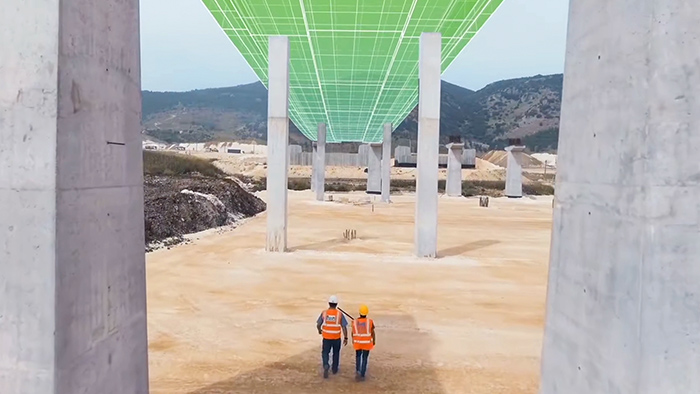 Deux personnes portant un gilet de sécurité orange et un casque de chantier sur un terrain aride marron avec des montagnes au loin, marchant sous une passerelle recouverte par une grille en vert et blanc