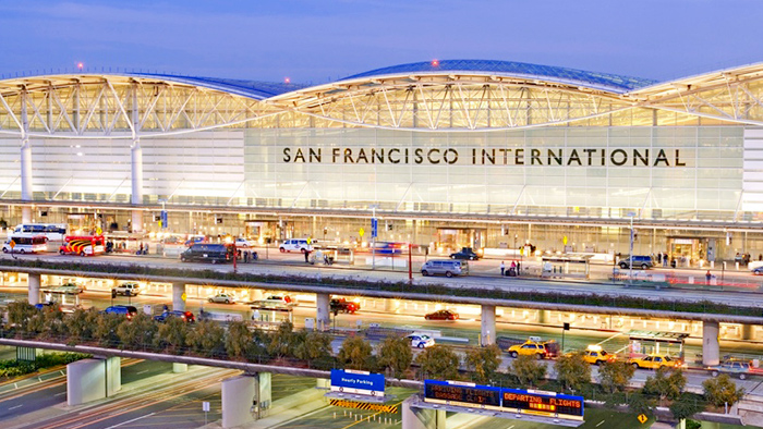 Façade de l’aéroport international de San Francisco devant laquelle passent plusieurs voitures sous un ciel bleu brumeux en soirée