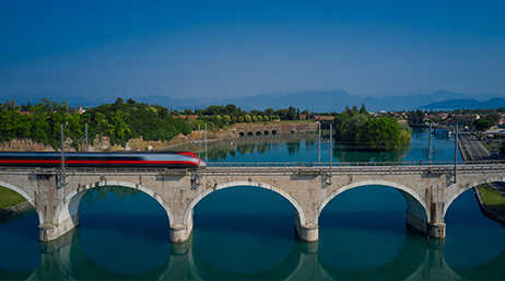 Train régional rouge et argent passant à grande vitesse sur un pont dont plusieurs arches enjambent une eau bleue calme sous un ciel bleu sans nuage