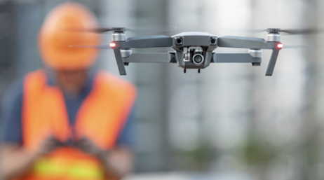 Drone en vol au premier plan avec un opérateur de terrain portant une veste et un casque de chantier orange en arrière-plan
