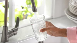 Main tenant un verre transparent sous un robinet d’eau en chrome dans une cuisine moderne blanche ensoleillée