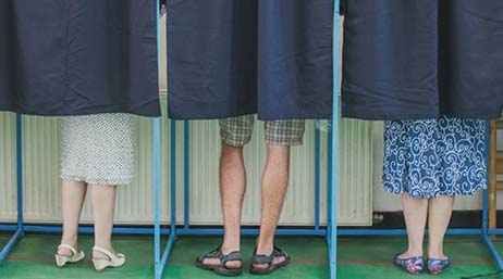 Photo d’une rangée d’isoloirs à rideau bleu ne laissant apparaître que les jambes des électeurs sous l’ourlet du rideau