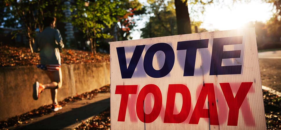 Une photo d'un panneau blanc avec écrit "VOTE TODAY" ("VOTEZ AUJOURD'HUI) en lettres bleues et rouges, se reposant sur un trottoir bordé d'arbres, alors qu'une personne en short et t-shirt court devant le lever du soleil dans le fond.