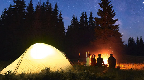 Groupe de personnes assises devant un feu de camp à la nuit tombée en forêt, avec leur tente éclairée
