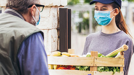 Photo de deux personnes en tenue décontractée se tenant à l’extérieur et portant un masque pendant que l’une d’entre elles tend à l’autre une caisse de produits colorés