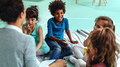 Photo de quatre enfants en train de rire et d’un adulte assis en cercle sur le sol d’une classe colorée