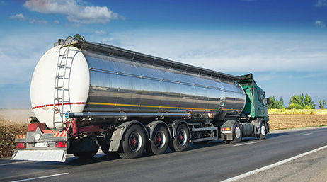 Long camion argent et vert sur une autoroute ensoleillée qui circule à travers des terres cultivées jaunes sous un ciel bleu vif