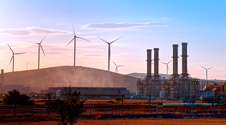 Complexe industriel à côté de plusieurs collines sur lesquelles se découpent la silhouette d’éoliennes sur fond de ciel bleu clair