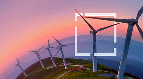 Versant de colline vert avec une rangée d’éoliennes blanches sur un horizon montagneux violet sous un ciel rose et orange au soleil couchant