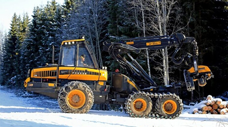 Photo d’un engin de travaux jaune et noir sur une route enneigée dans une forêt hivernale à côté d’une pile de rondins