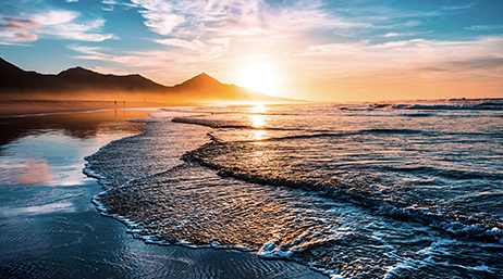 Vagues peu profondes frappant doucement le rivage avec des montagnes au loin et le soleil à l’horizon