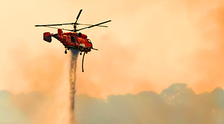 Hélicoptère rouge lâchant un produit ignifuge sur un brasier dans un ciel enfumé de teinte orange