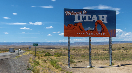 Portion d'autoroute nationale dans un vaste désert couvert de broussailles avec un grand panneau coloré indiquant "Bienvenue dans l'Utah", sous un ciel bleu vif