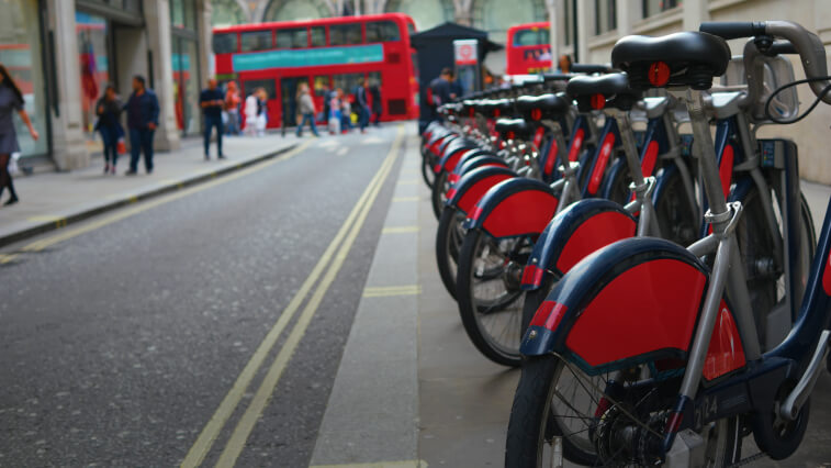 Un service de vélos en libre-service au bord d’une rue, avec des piétons et des bus à impériale au loin