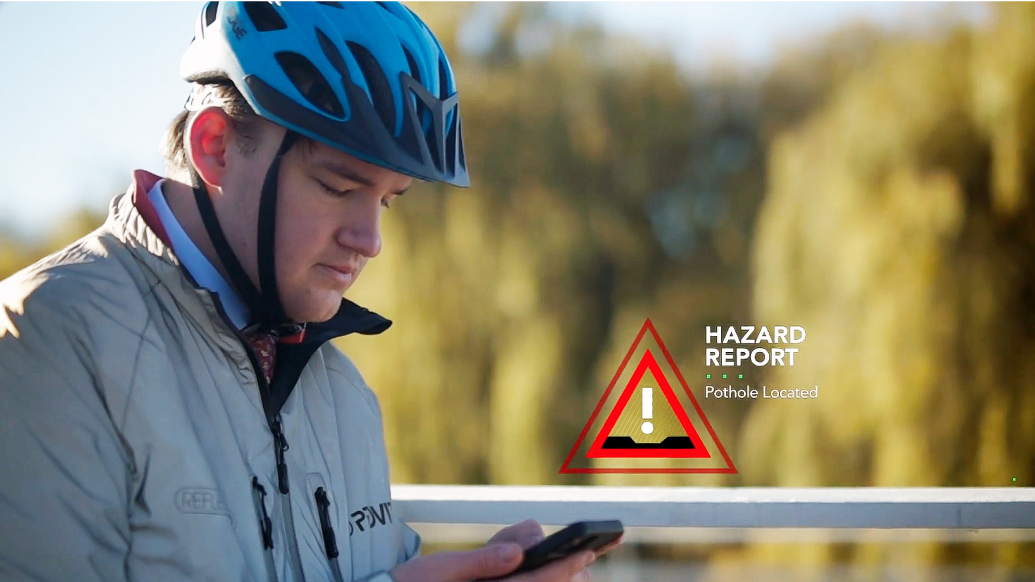 Une personne en coupe-vent et casque de vélo consulte un téléphone portable, avec des arbres en arrière-plan.